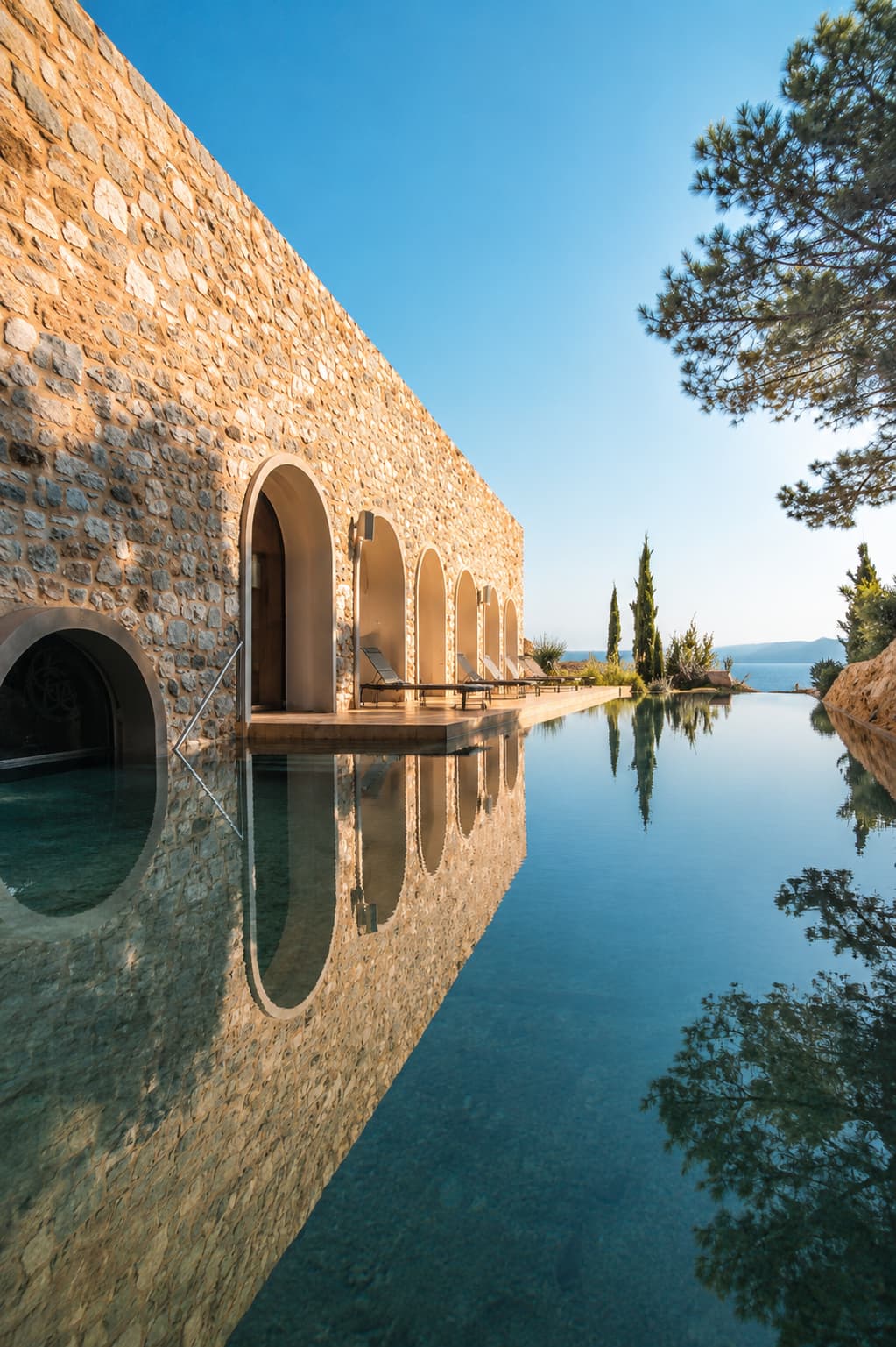 Stone spa building and still infinity pool framed by cypress trees at a solo-friendly wellness retreat in Greece