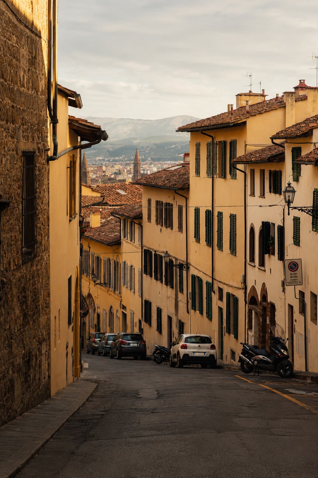 A quiet sloping street in the Oltrarno, shuttered ochre houses lining the hill with Florence's rooftops and distant mountains beyond