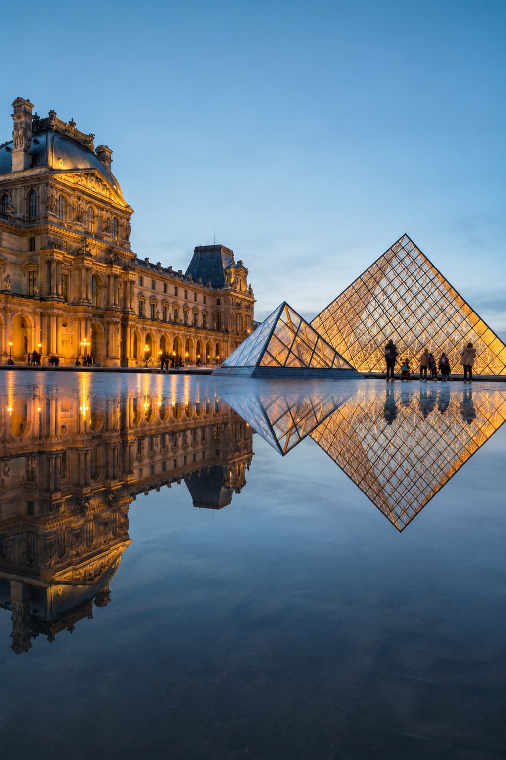 The Louvre Pyramid lit up at dusk, reflected in the wet courtyard pavement