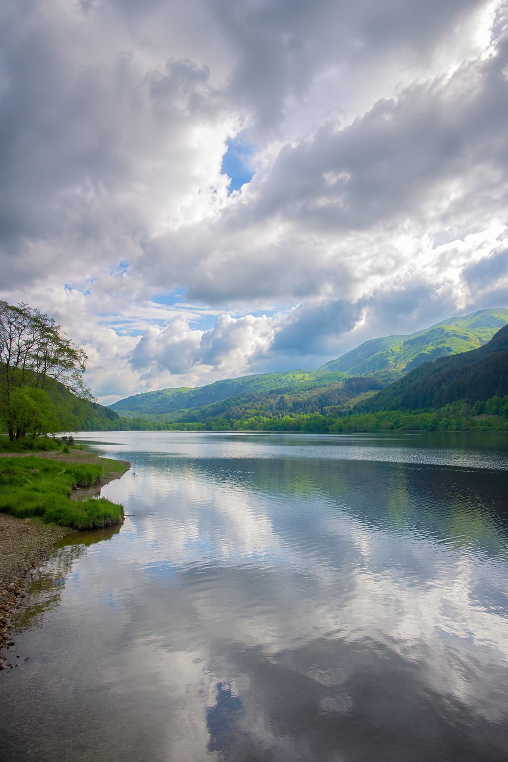 A still loch reflecting clouds and green hills in the Scottish Highlands on a bright spring day
