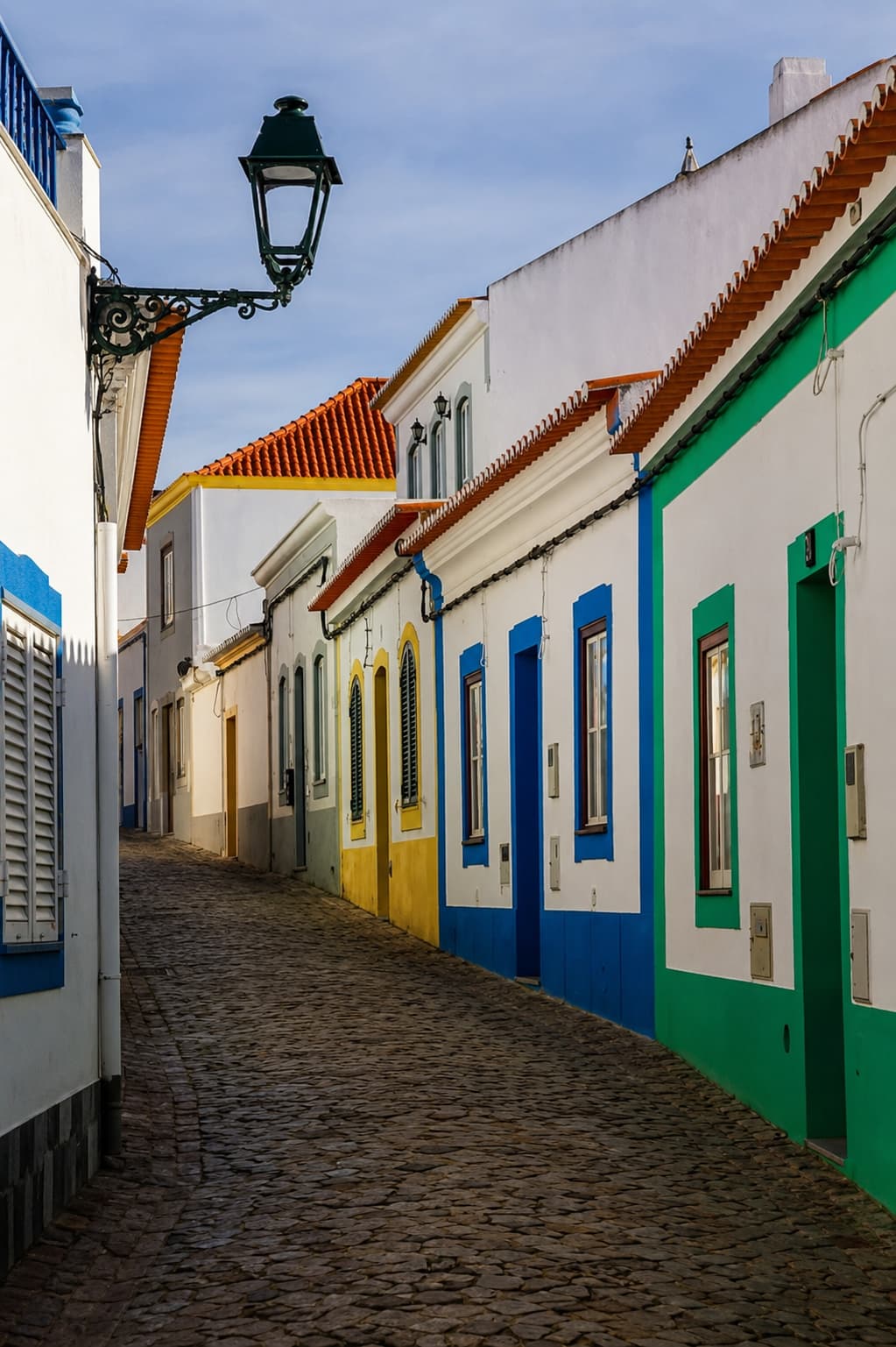 Empty cobblestone street in a traditional Algarve village with whitewashed houses painted in yellow, blue, and green, and a wrought-iron street lamp overhead