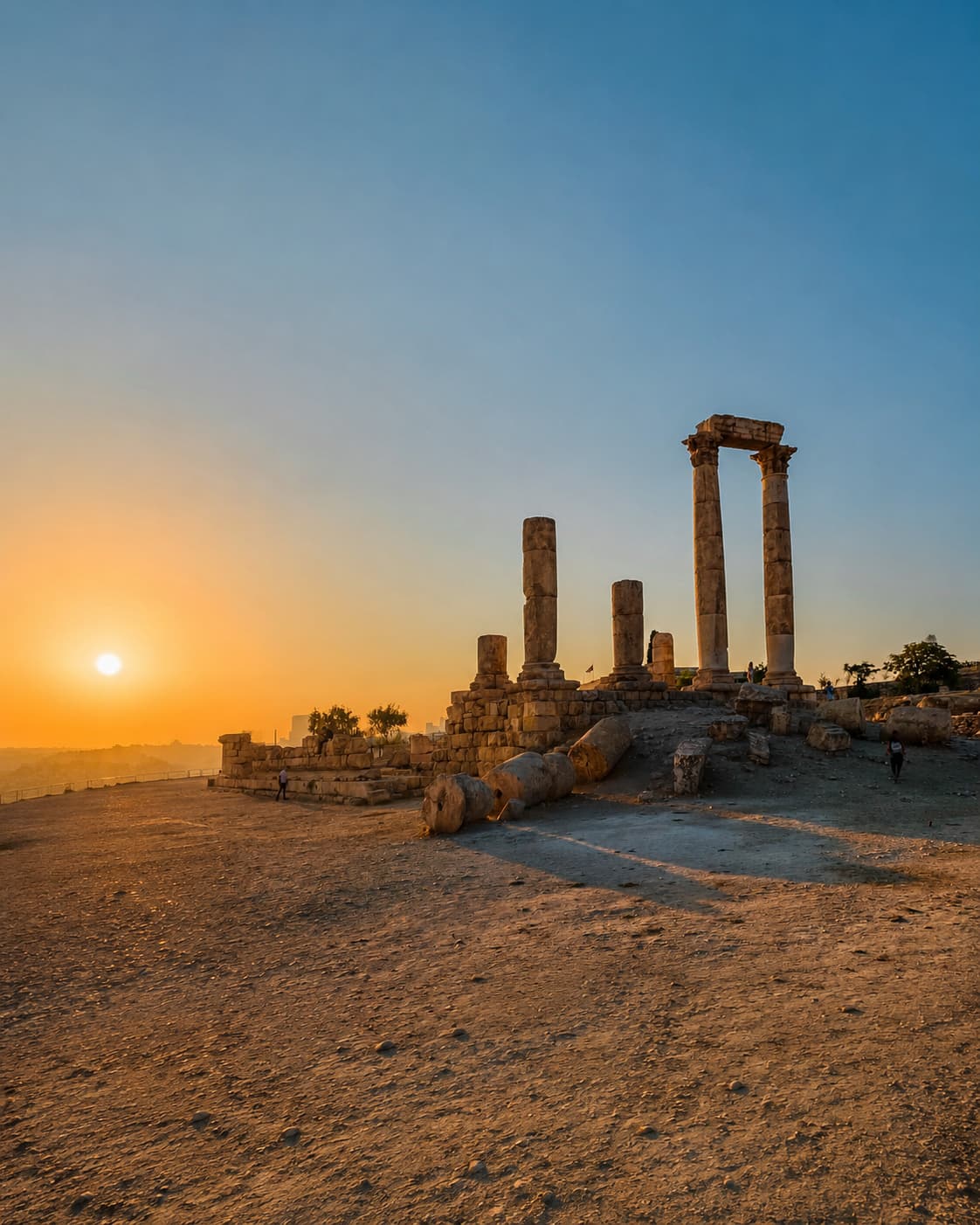 The standing columns of the Temple of Hercules at the Amman Citadel silhouetted against an orange sunset over the hills