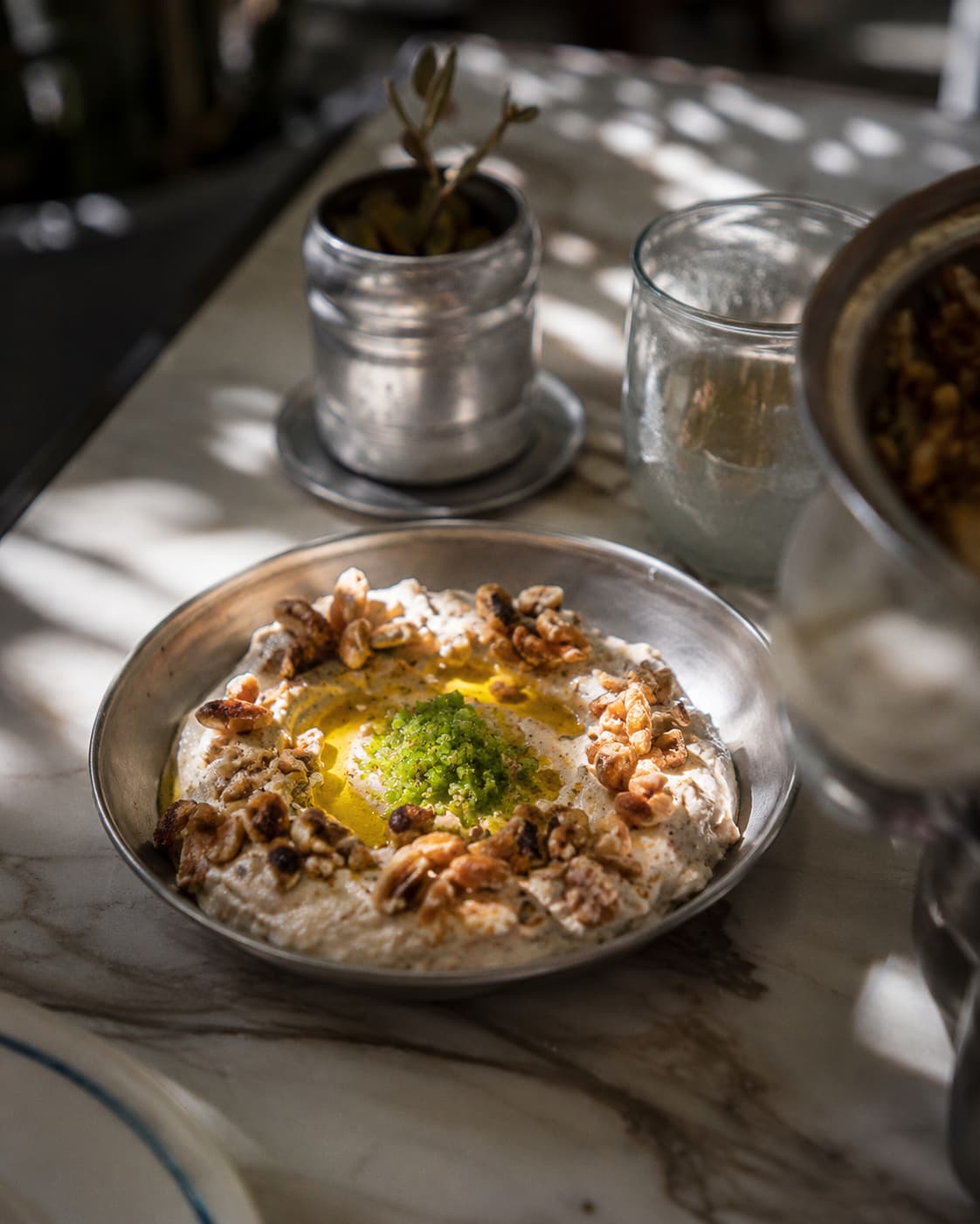 A bowl of Jordanian mezze — a creamy dip topped with olive oil, toasted walnuts and a spoon of green herb salsa — on a sunlit marble table