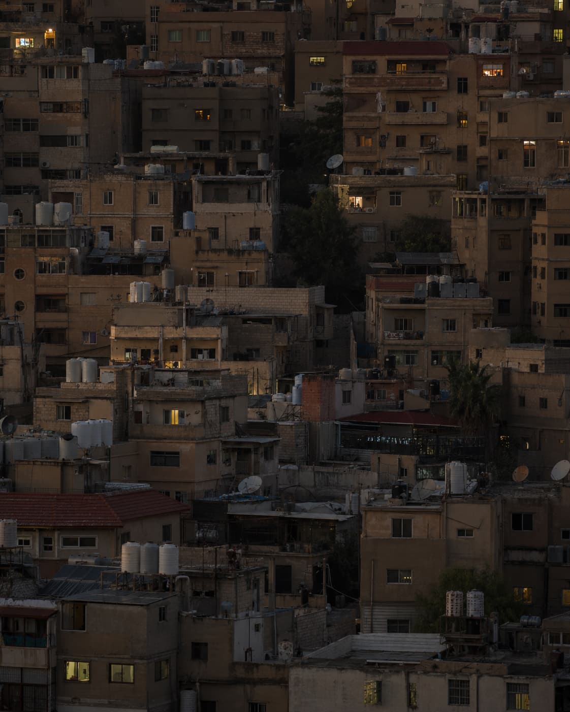 Amman's stacked hillside of pale stone buildings at dusk, warm window lights coming on across the seven hills of the city