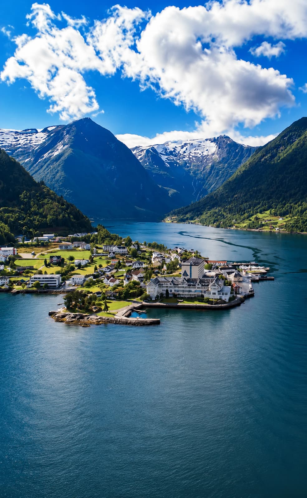 A small Norwegian fjord village on a green spit of land, snow-streaked mountains rising behind deep blue water in summer