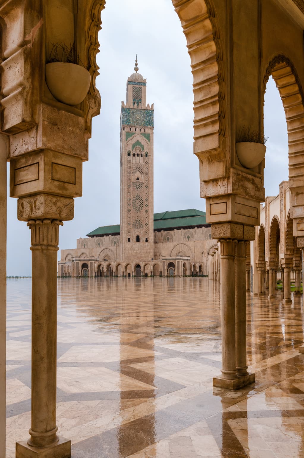 Hassan II Mosque and its tiled minaret seen through carved marble arches across the rain-polished courtyard in Casablanca