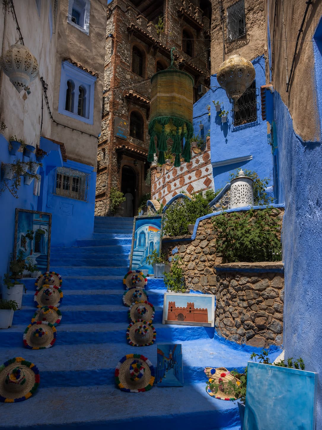 A blue-painted stairway in Chefchaouen's medina lined with colourful straw hats and small paintings for sale