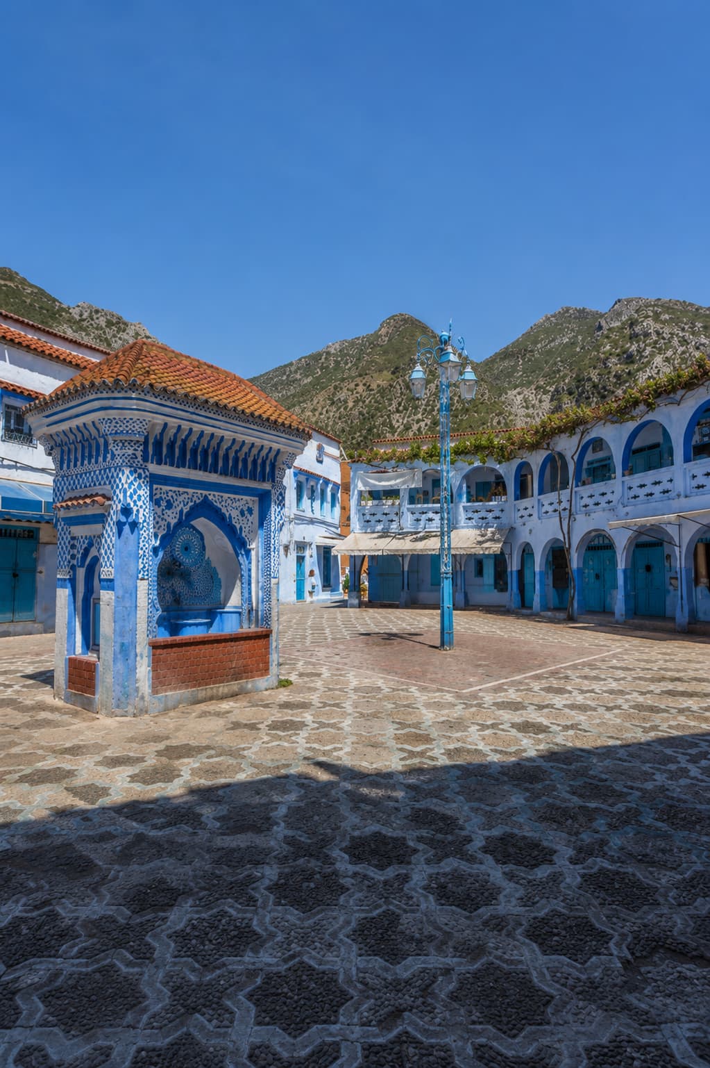 Place Outa el-Hammam, the central square of Chefchaouen, with a blue-and-white fountain, Islamic arches, and the Rif Mountains behind