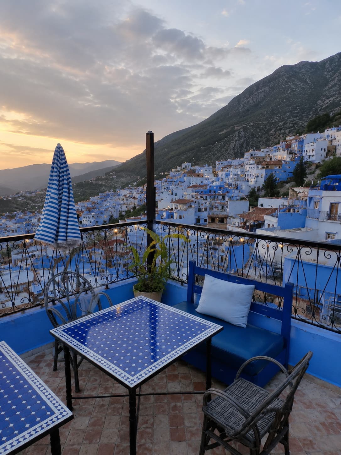 A riad rooftop terrace in Chefchaouen at dusk, with blue mosaic tables and chairs overlooking the blue medina and Rif Mountains