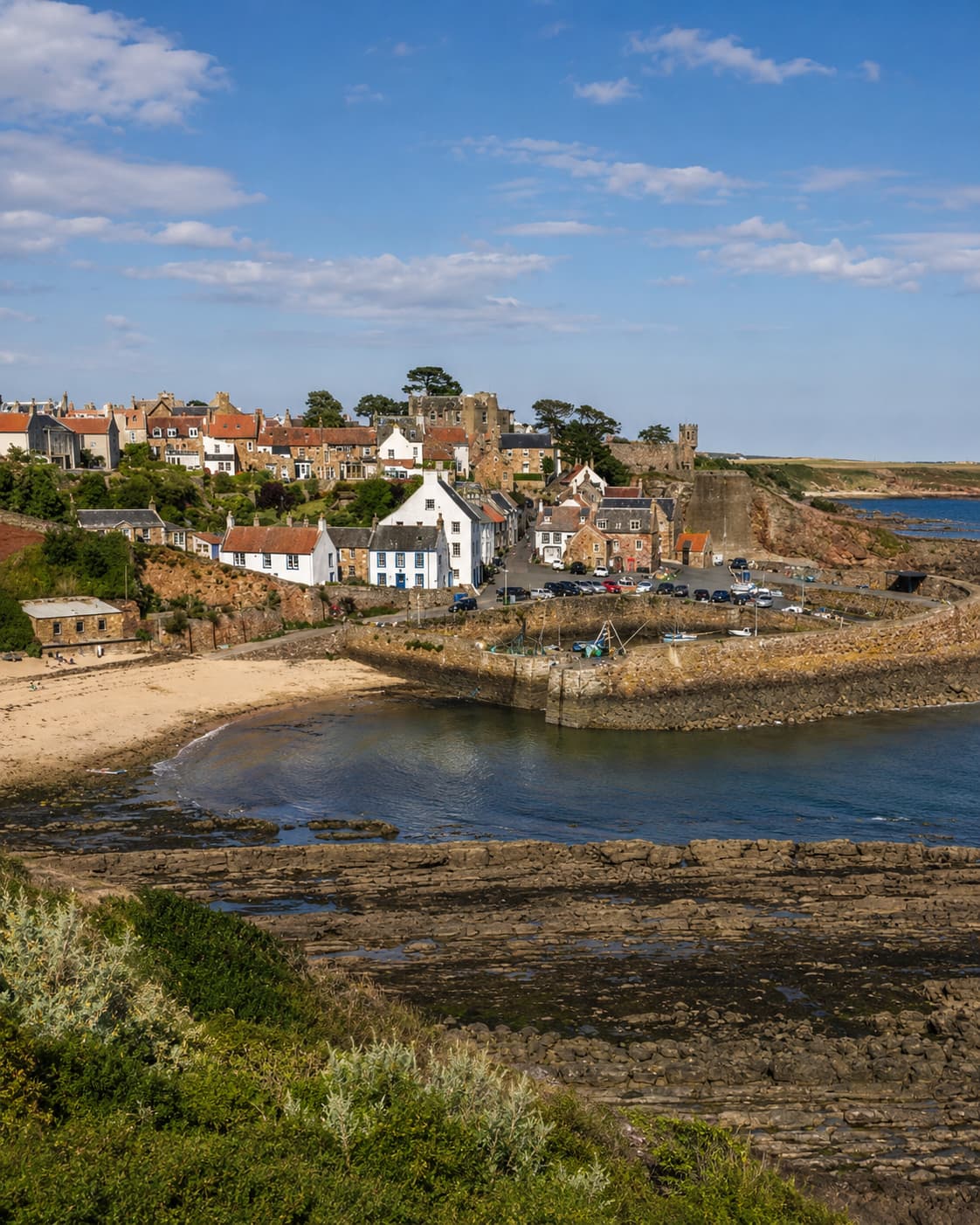Crail harbour East Neuk of Fife Scotland — stone harbour walls, sandy beach, whitewashed and stone cottages on the hillside above, blue sky, most beautiful coastal village in Scotland