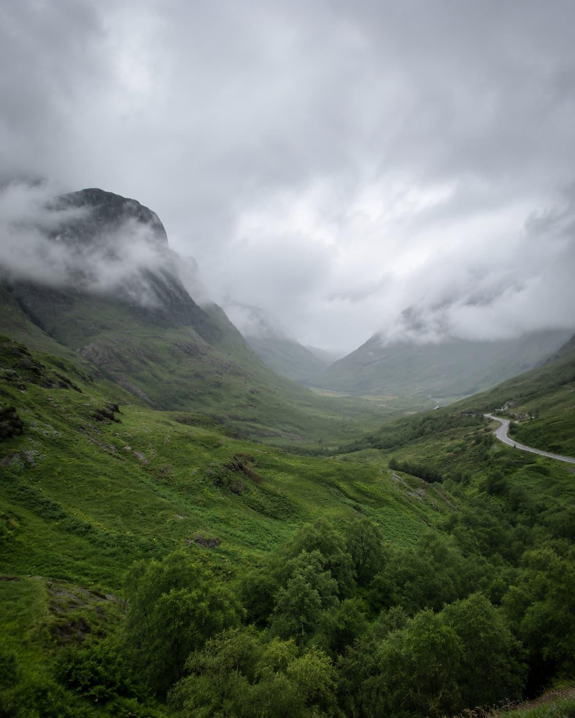 Glencoe valley Scotland — dramatic green mountains shrouded in low cloud and mist, winding road through the valley, one of the most atmospheric Highland villages in Scotland