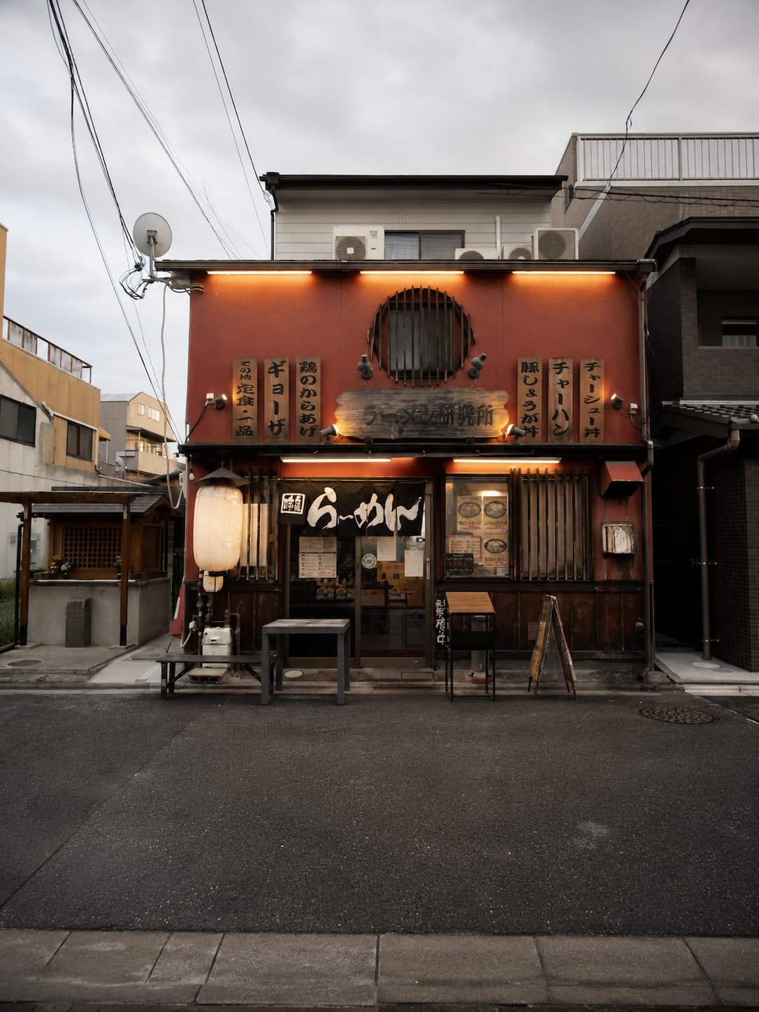 japan solo travel - A traditional Japanese ramen shop facade at dusk — warm orange lantern light, hand-painted kanji signs, and an empty bench outside