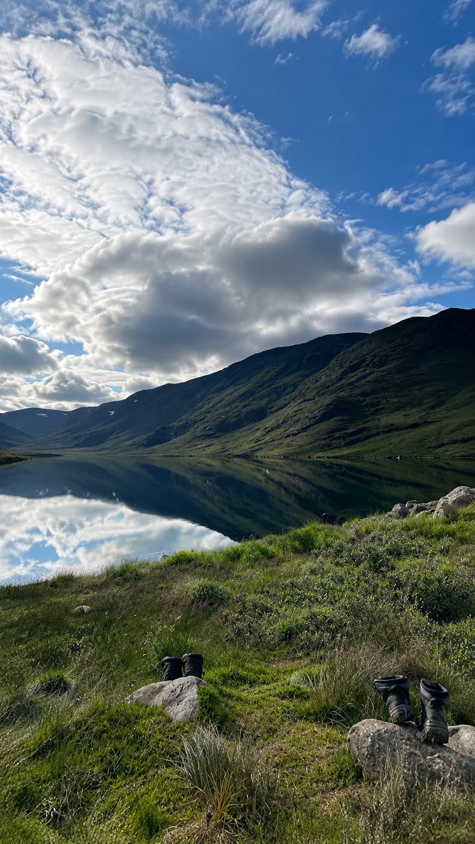 Hiking boots resting beside a glassy mountain lake in Swedish Lapland in July, with a perfect cloud reflection on still water — July travel in the far north at its best
