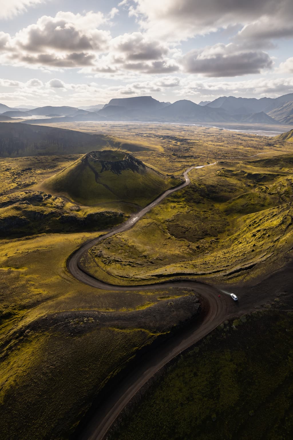 Aerial view of a winding gravel F-road through Iceland's green volcanic highland interior with a white campervan — July is the only month these roads are fully open