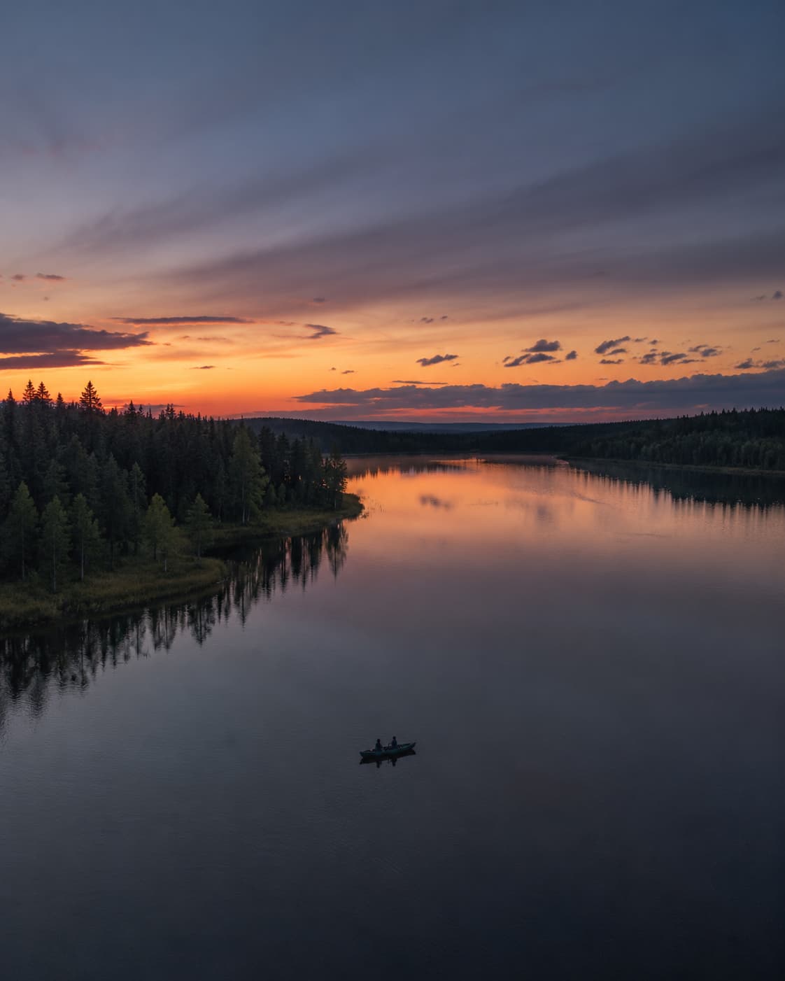 A lone rowing boat silhouetted on a glassy Swedish Lapland lake at midnight sun, with pine forest reflections and an orange and purple twilight sky — one of the best places to go in July for light