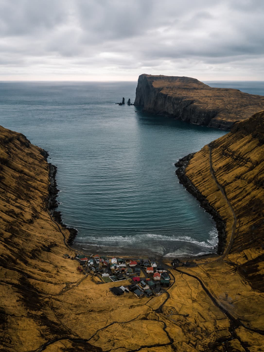 Aerial view of a remote Faroese village at the foot of dramatic ocean cliffs with a sea stack in the distance — the Faroe Islands in July at the peak of their green season