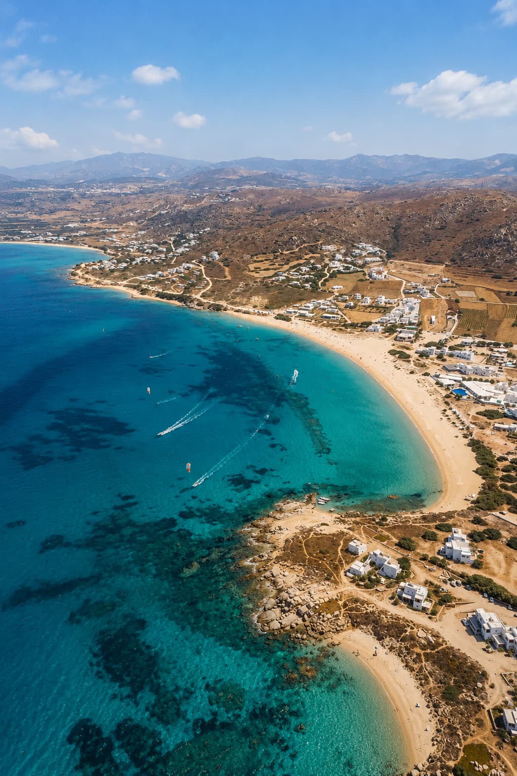 Aerial view of a Greek island beach with turquoise and blue Aegean water, white Cycladic buildings, and kitesurfers in July before the August peak crowd season