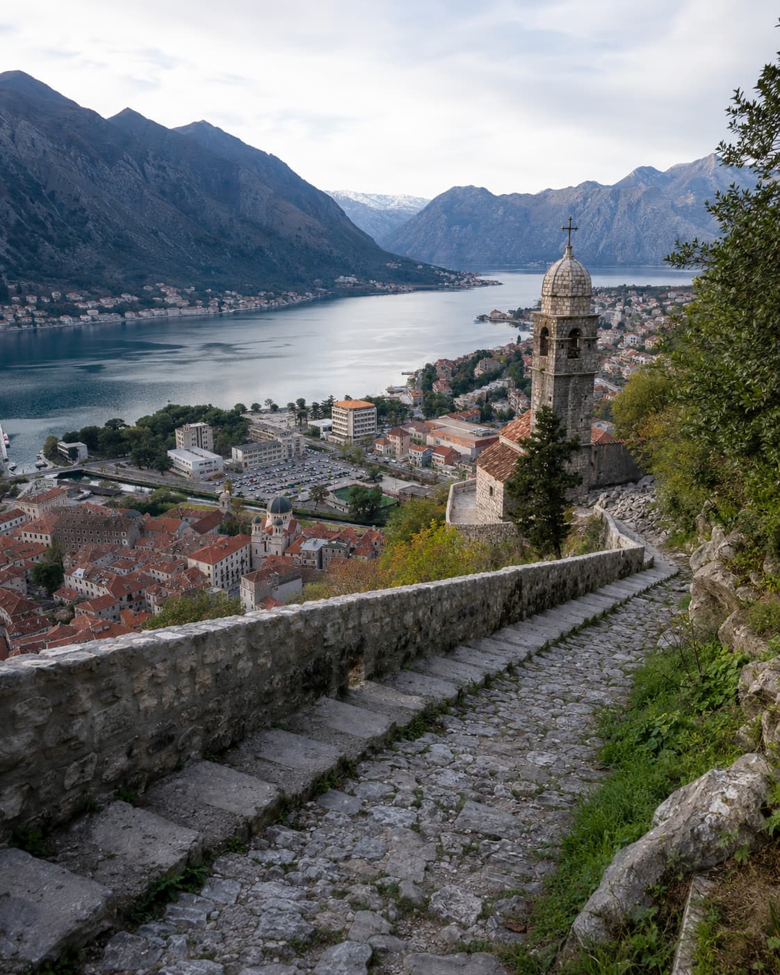 Stone stairway climbing the fortress walls above Kotor old town in Montenegro, with the bay and snow-capped mountains beyond — one of the best July travel destinations on the Adriatic