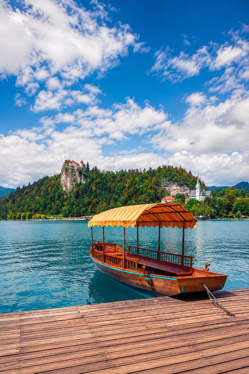 Traditional wooden pletna boat moored at Lake Bled dock with Bled Island church and clifftop Bled Castle behind the turquoise water — the midday highlight of any Lake Bled autumn itinerary