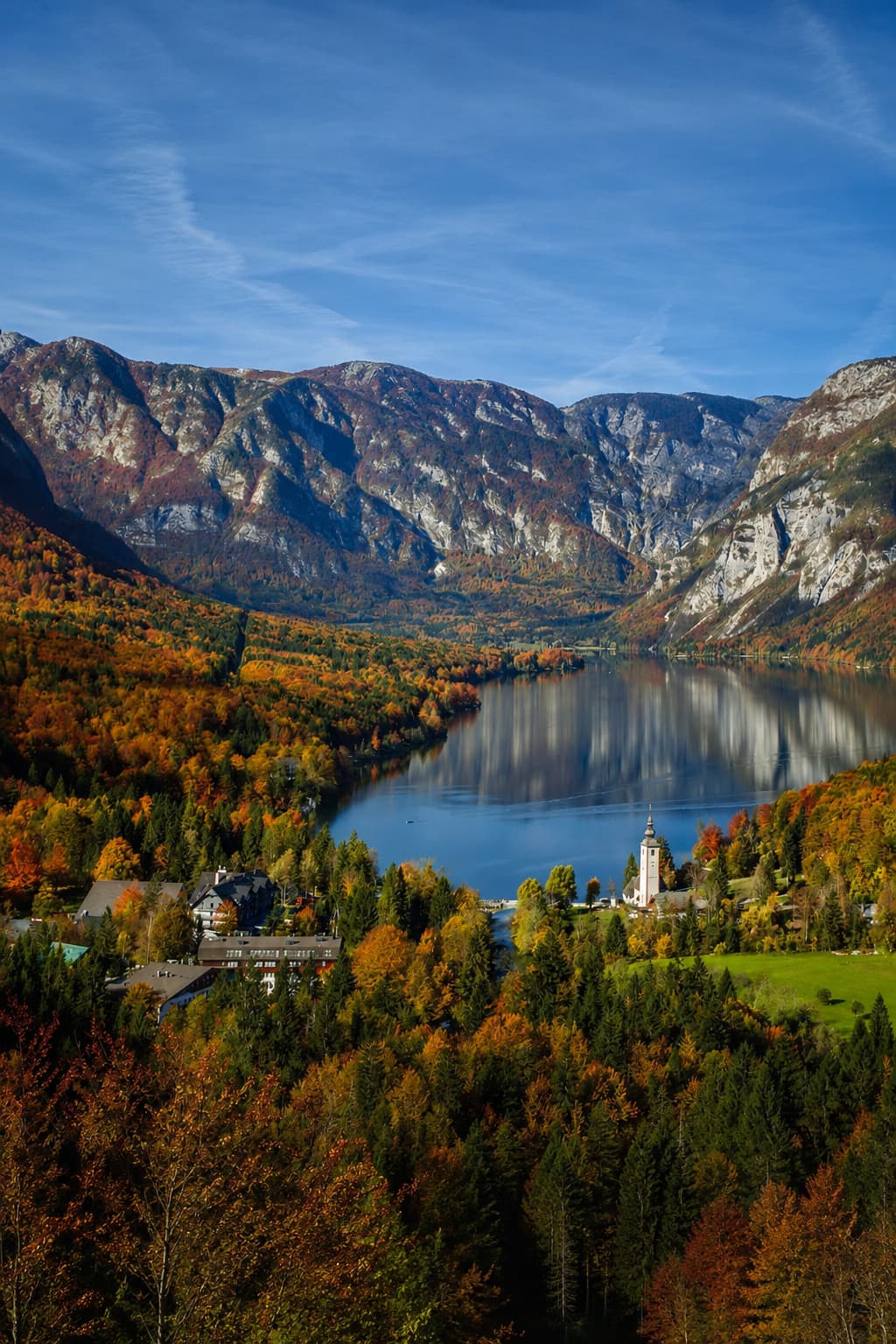 Lake Bohinj in peak autumn colour on a Lake Bled autumn itinerary day trip — red and orange forested slopes reflected in the still blue water, Julian Alps rising steeply above, Slovenia