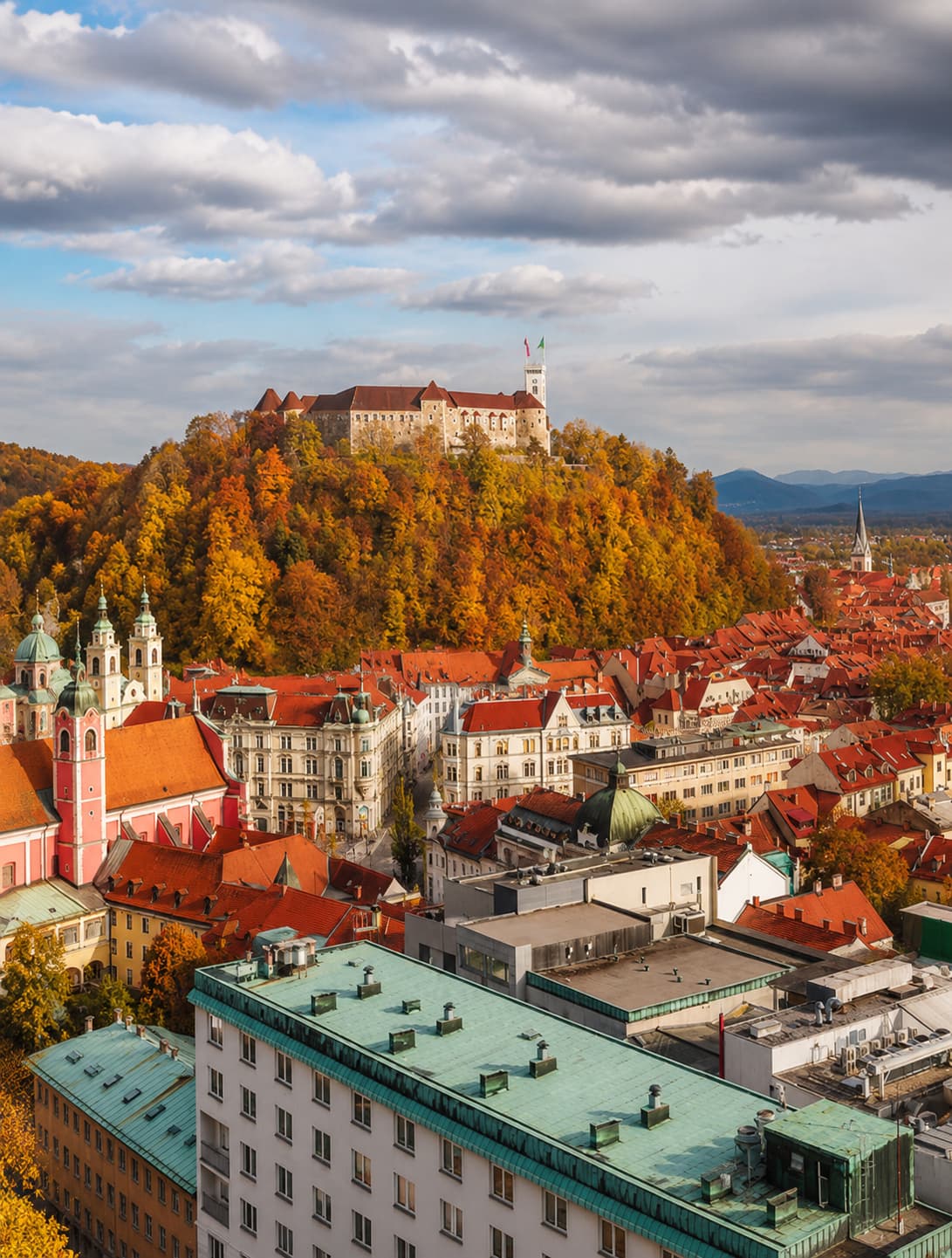 Ljubljana Castle perched on a forested hill surrounded by gold and orange autumn canopy, rising above the old town's red rooftops