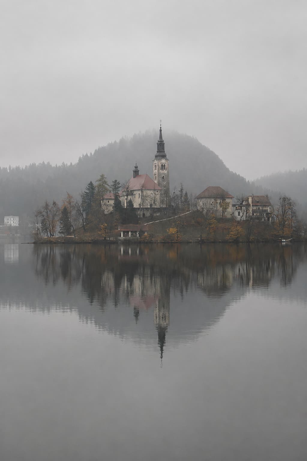 Lake Bled island and church reflected in perfectly still grey water on a heavy fog morning in late autumn