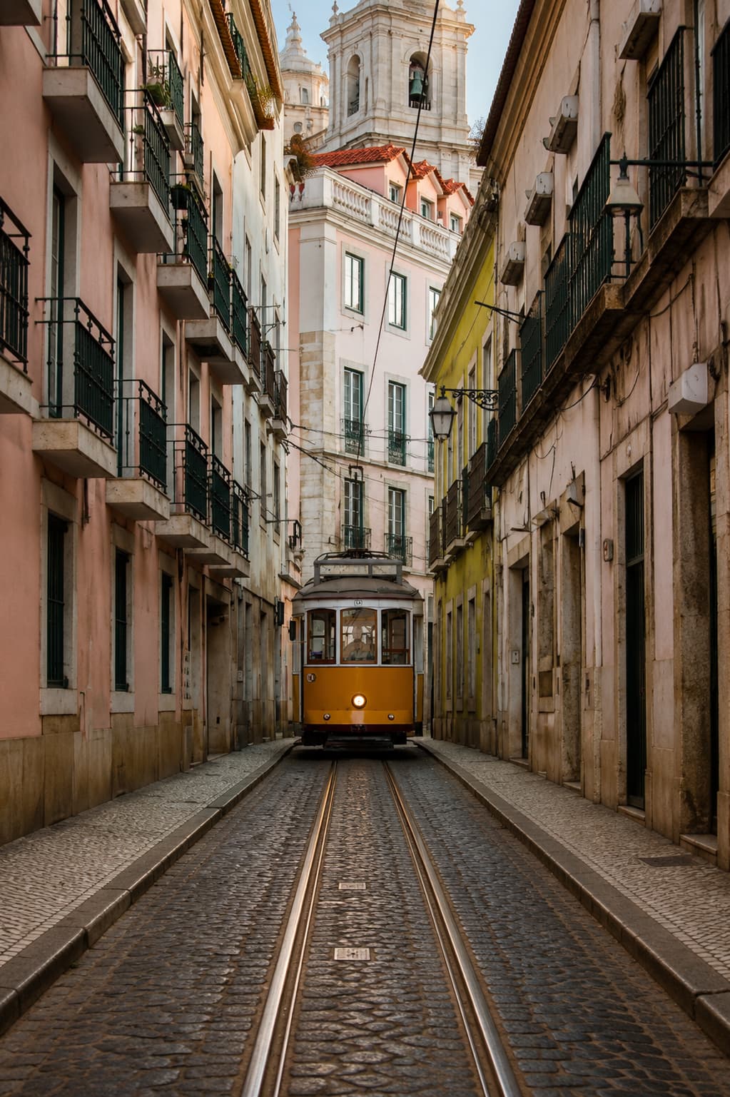 Yellow tram on a narrow cobblestone street in Lisbon heading toward a church tower