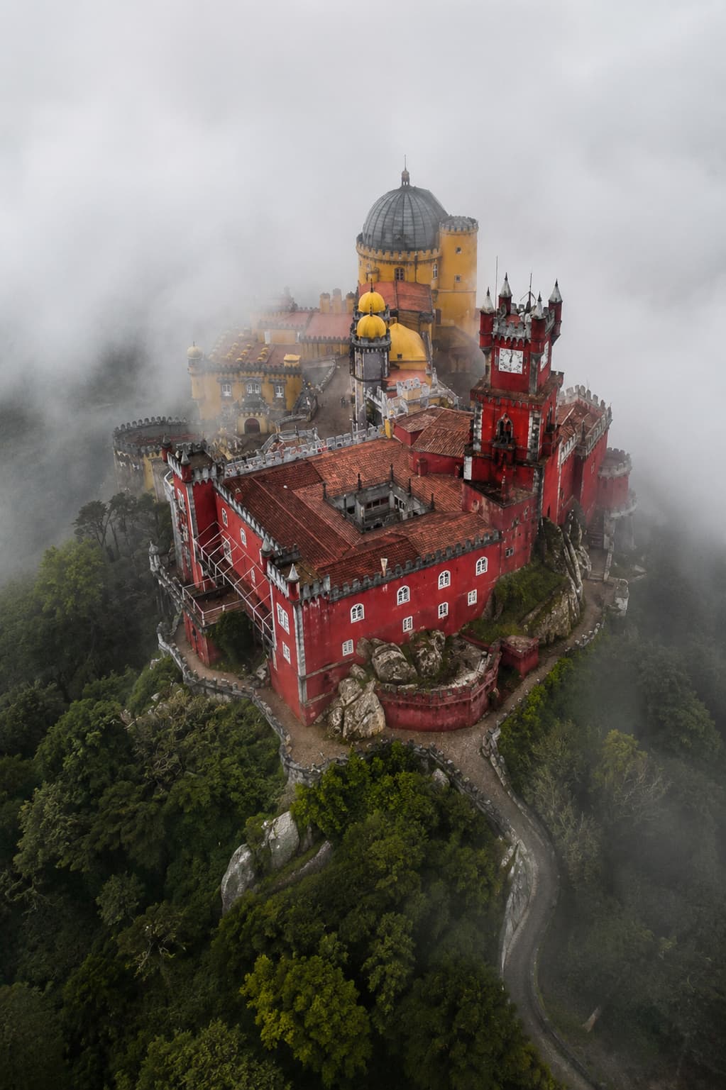 Aerial view of Pena Palace in Sintra emerging from low cloud, red and yellow towers above forested hills