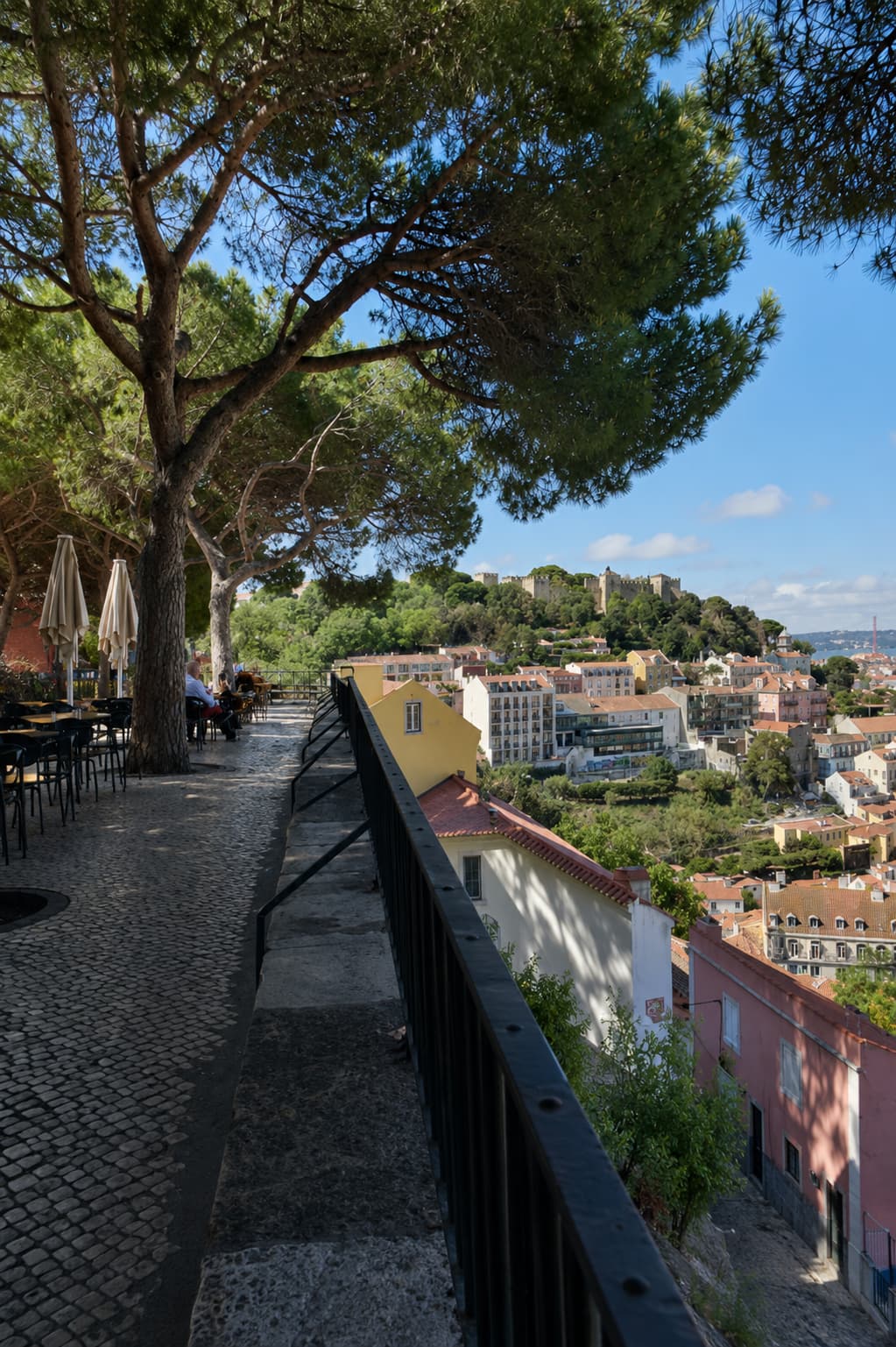 Miradouro da Graça viewpoint in Lisbon with pine tree canopy, café terrace, and São Jorge Castle visible across the rooftops
