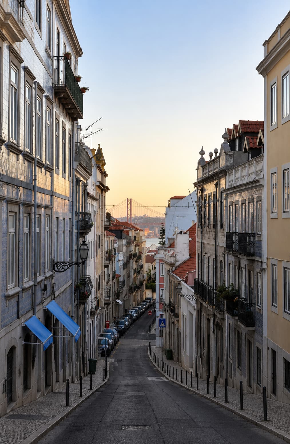 Steep residential street in Lisbon at sunset with azulejo-tiled buildings and the 25 de Abril bridge visible in the distance