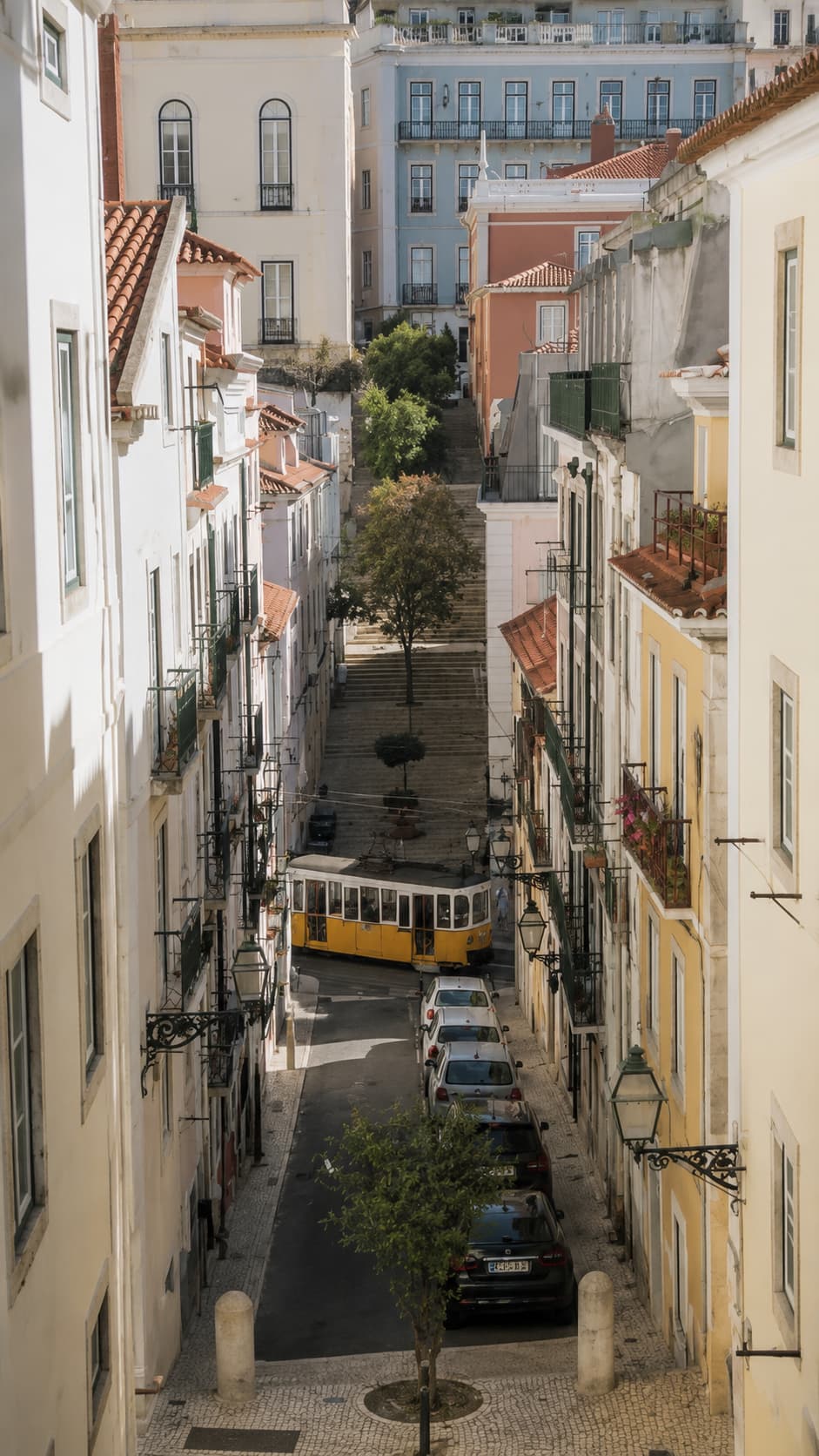 Aerial view down a narrow Lisbon street with a yellow tram visible below between pastel buildings and stone steps