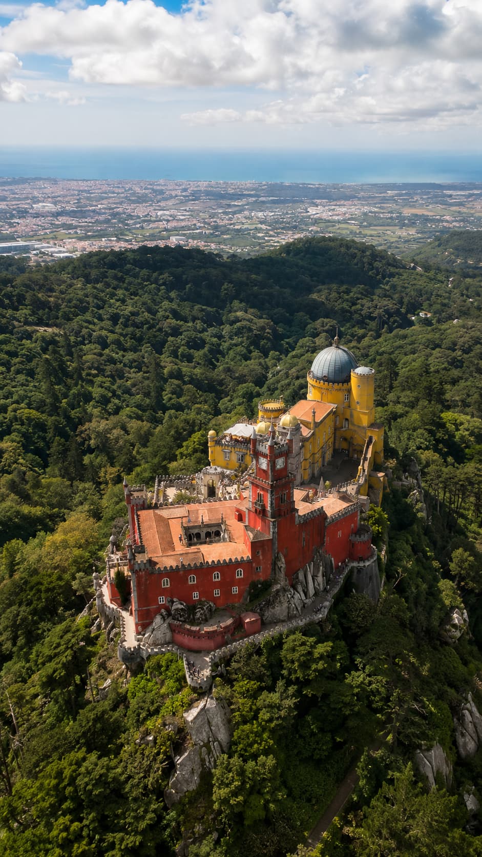 Aerial view of Pena Palace in Sintra — red and yellow palace perched on a forested hilltop with the Atlantic coastline visible in the distance
