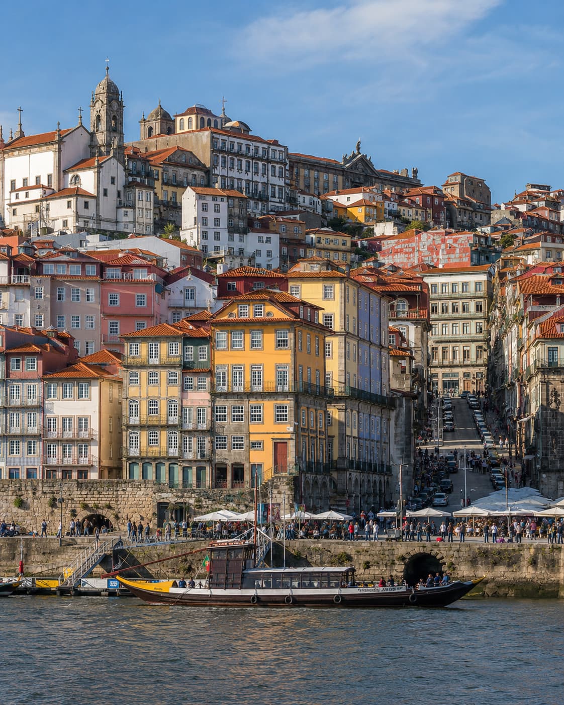 Porto's Ribeira waterfront viewed from across the Douro — colourful stacked facades climbing the hillside, rabelo boats moored at the quay