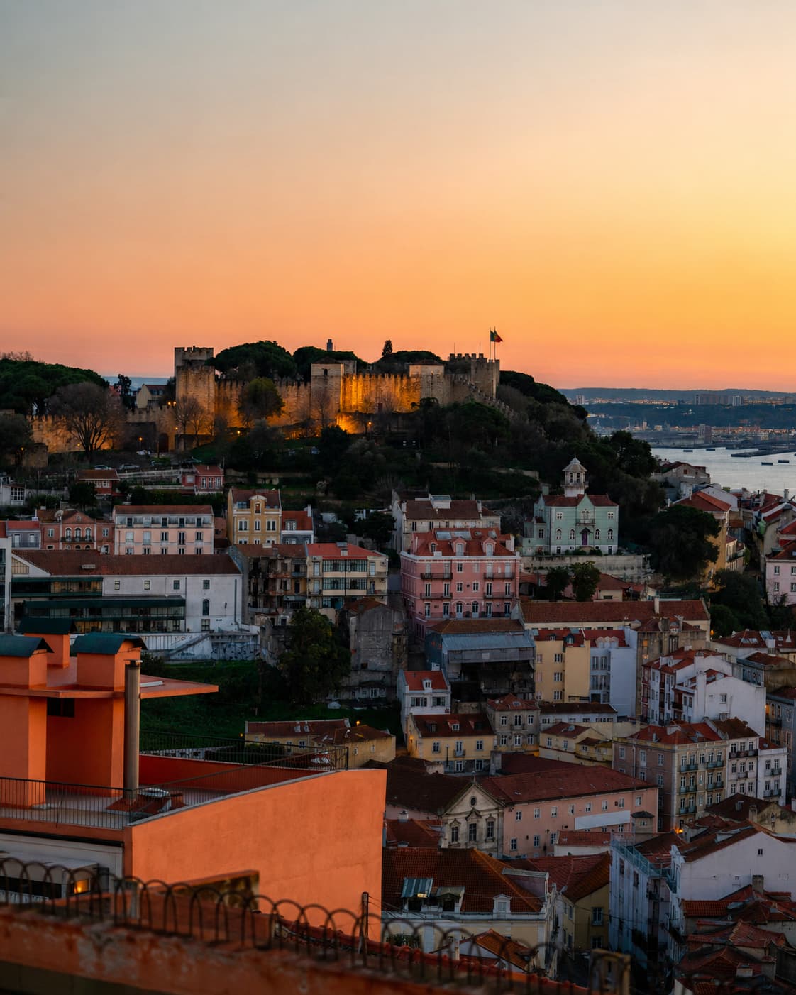 Lisbon at sunset — São Jorge Castle lit up gold on the hilltop above the rooftops, orange sky fading to the Tagus river below