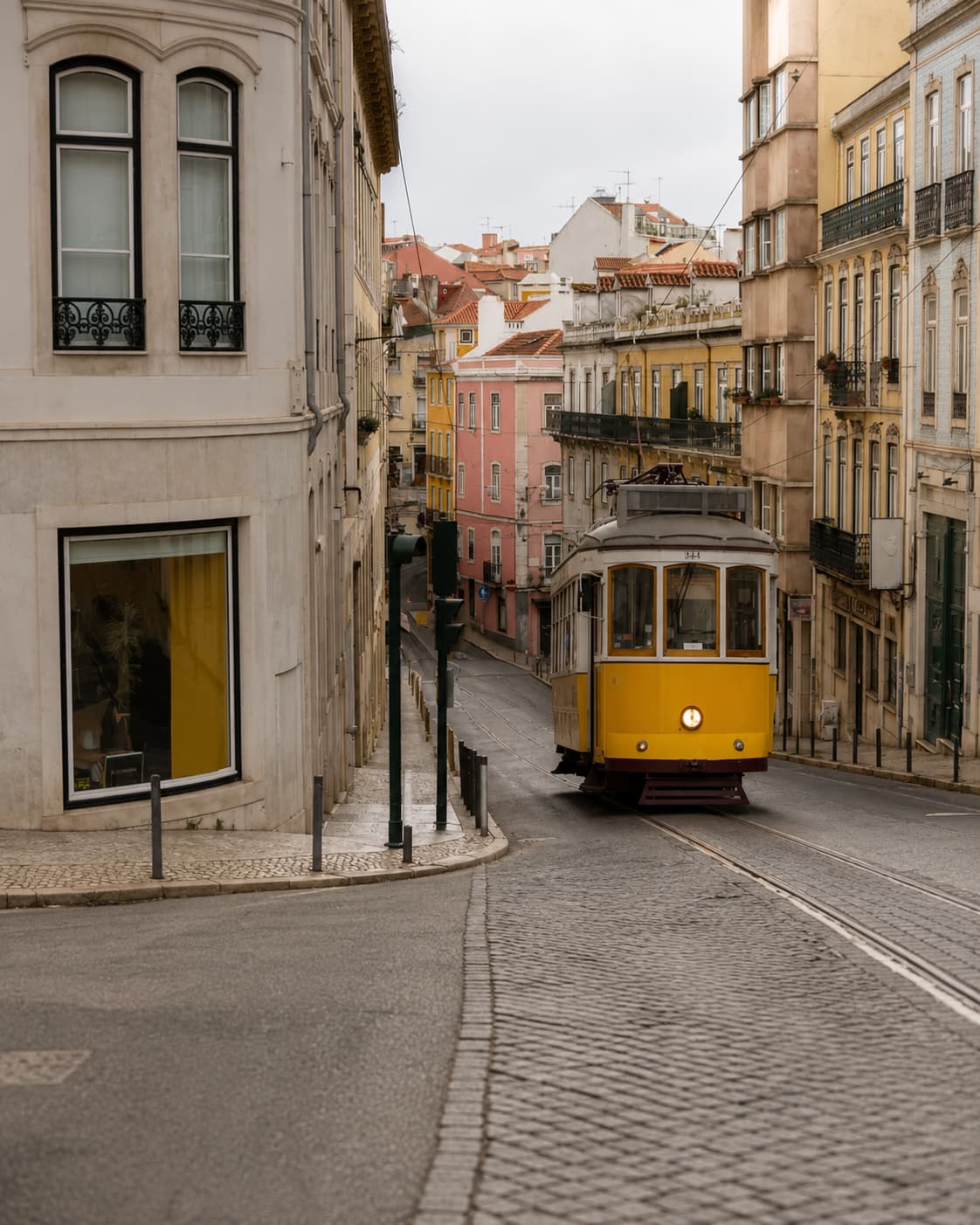 Lisbon's iconic yellow tram rounding a bend on a quiet cobbled street, pink and yellow building facades lining the hill behind
