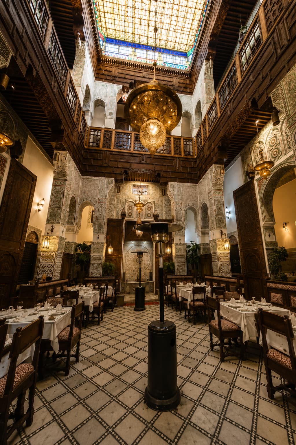 Grand riad dining room in Fes — ornate carved plaster walls, brass chandelier, stained-glass skylight, and white-clothed tables set for dinner on a zellige-tiled floor