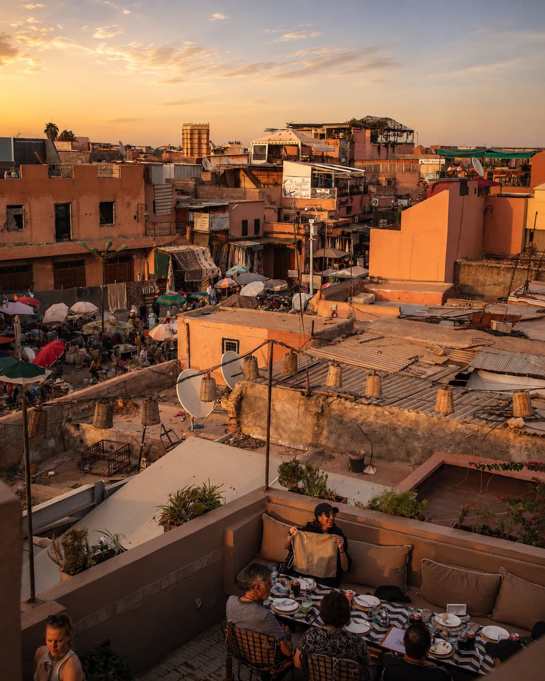 Rooftop dinner at sunset over the Marrakech medina — diners at a terrace table with ochre rooftops, satellite dishes, and a golden sky stretching behind them
