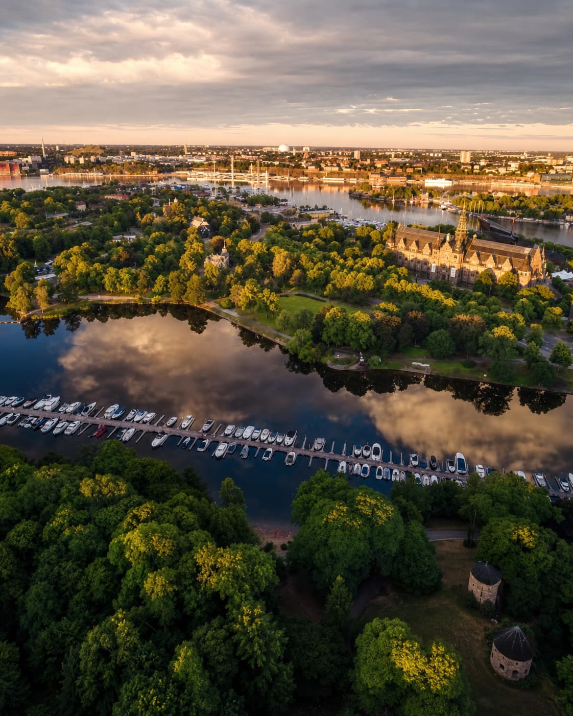 Aerial view of Stockholm's Djurgården island at golden hour — green tree canopy surrounding the Nordic Museum's ornate red-brick facade, boats moored along the waterway, and the city spreading to the horizon under a warm evening sky, showing Stockholm's distinctive summer character that makes it one of Europe's best August city breaks.