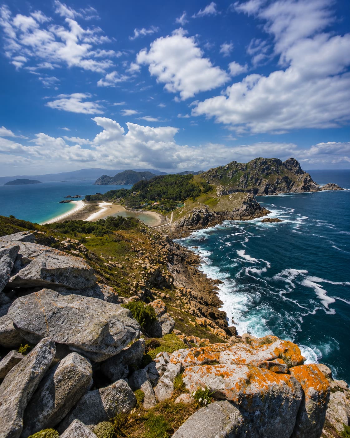 The Cíes Islands off the coast of Galicia in northwestern Spain — rocky coastline with crashing Atlantic surf in deep teal water, green hillsides and granite boulders in the foreground, and a sheltered white sand beach visible in the distance, capturing the wild Atlantic character that makes northern Spain a genuine August alternative to the Mediterranean heat.