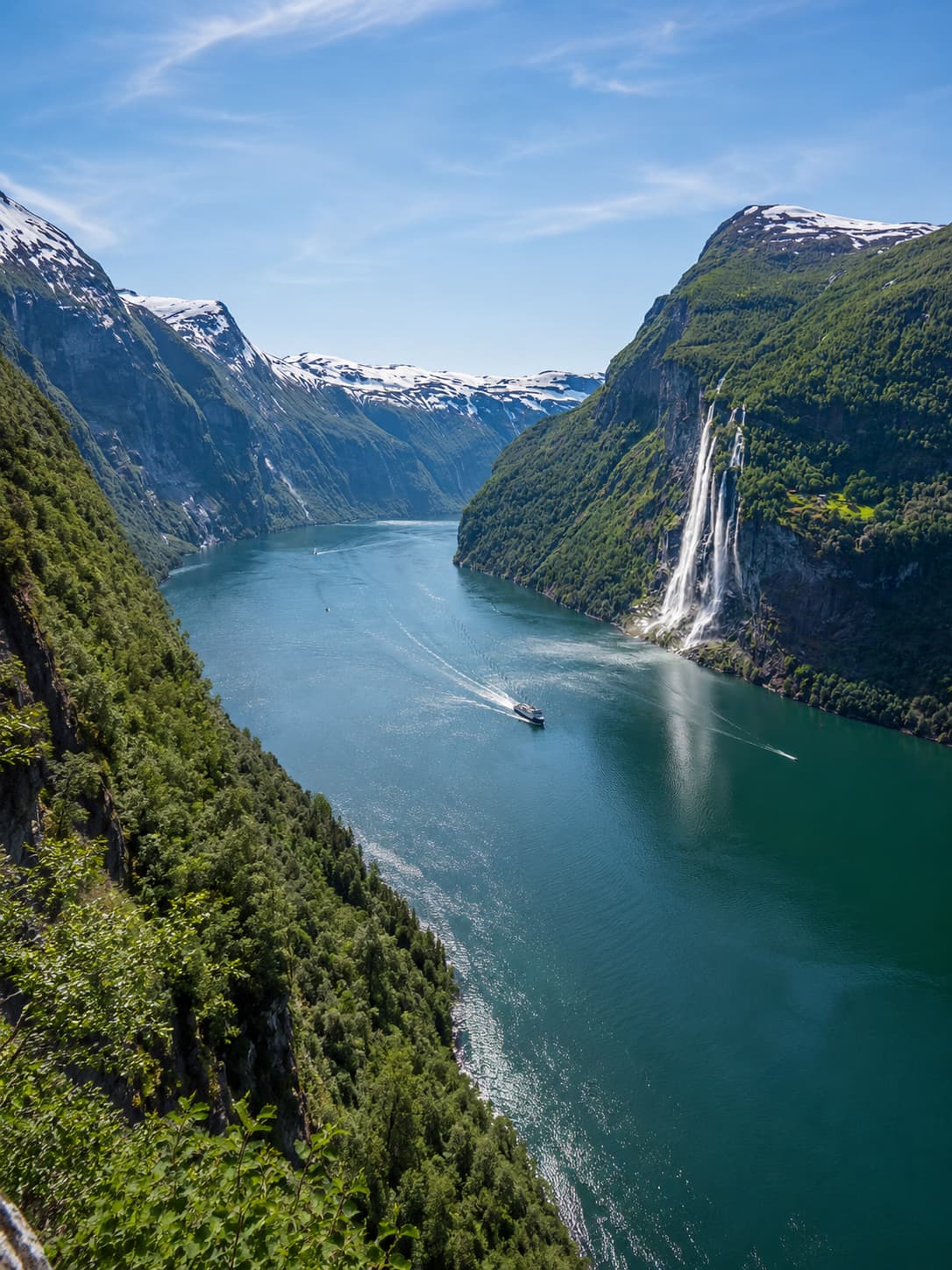 The Geirangerfjord in Norway — a deep green fjord winding between steep forested mountains with snow still on the highest peaks, a ferry crossing the still water and the Seven Sisters waterfall cascading down the cliff face, the quintessential image of cool, dramatic northern Europe that awaits travellers who decide the Mediterranean is too hot in August.