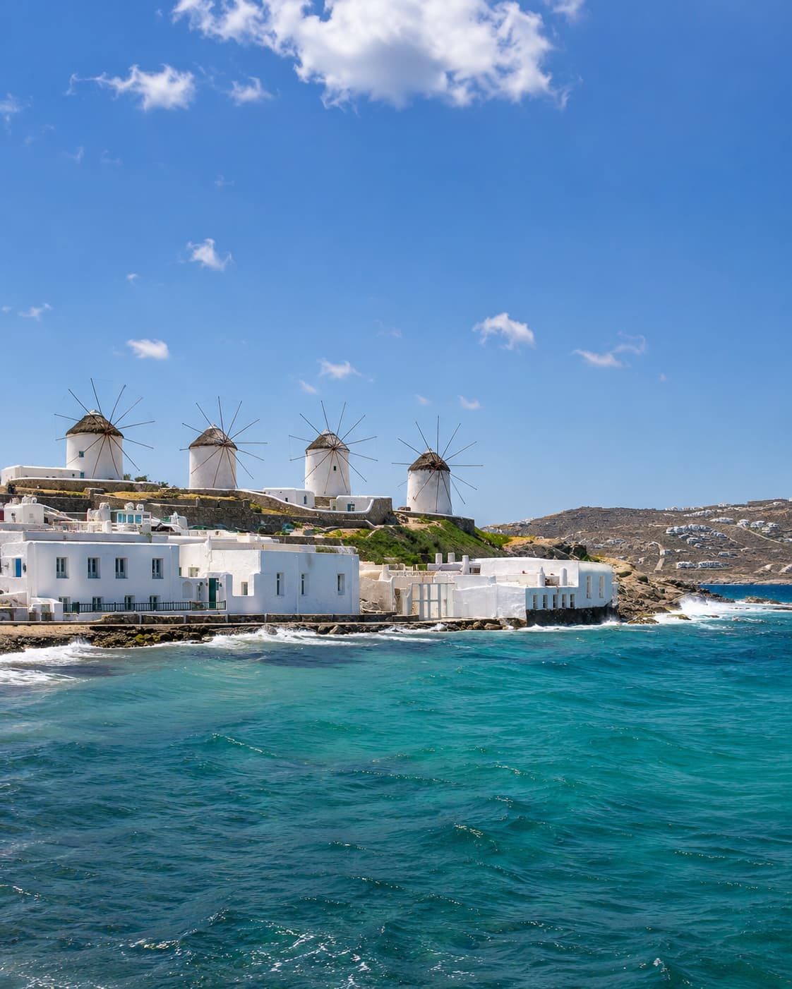 The iconic windmills of Mykonos on a hillside above the Aegean Sea — white Cycladic architecture against vivid turquoise water under a bright summer sky, representing the part of the Mediterranean that still works in August thanks to the cooling Meltemi wind.