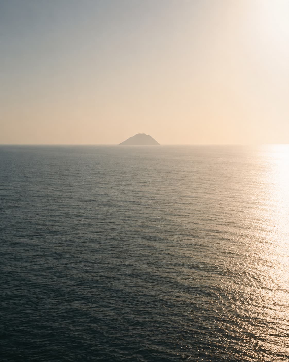 A lone volcanic island silhouetted against a hazy, sun-bleached Mediterranean sky — Stromboli rising from a glassy sea with atmospheric haze softening the horizon, capturing the quiet intensity of the Mediterranean in peak summer.