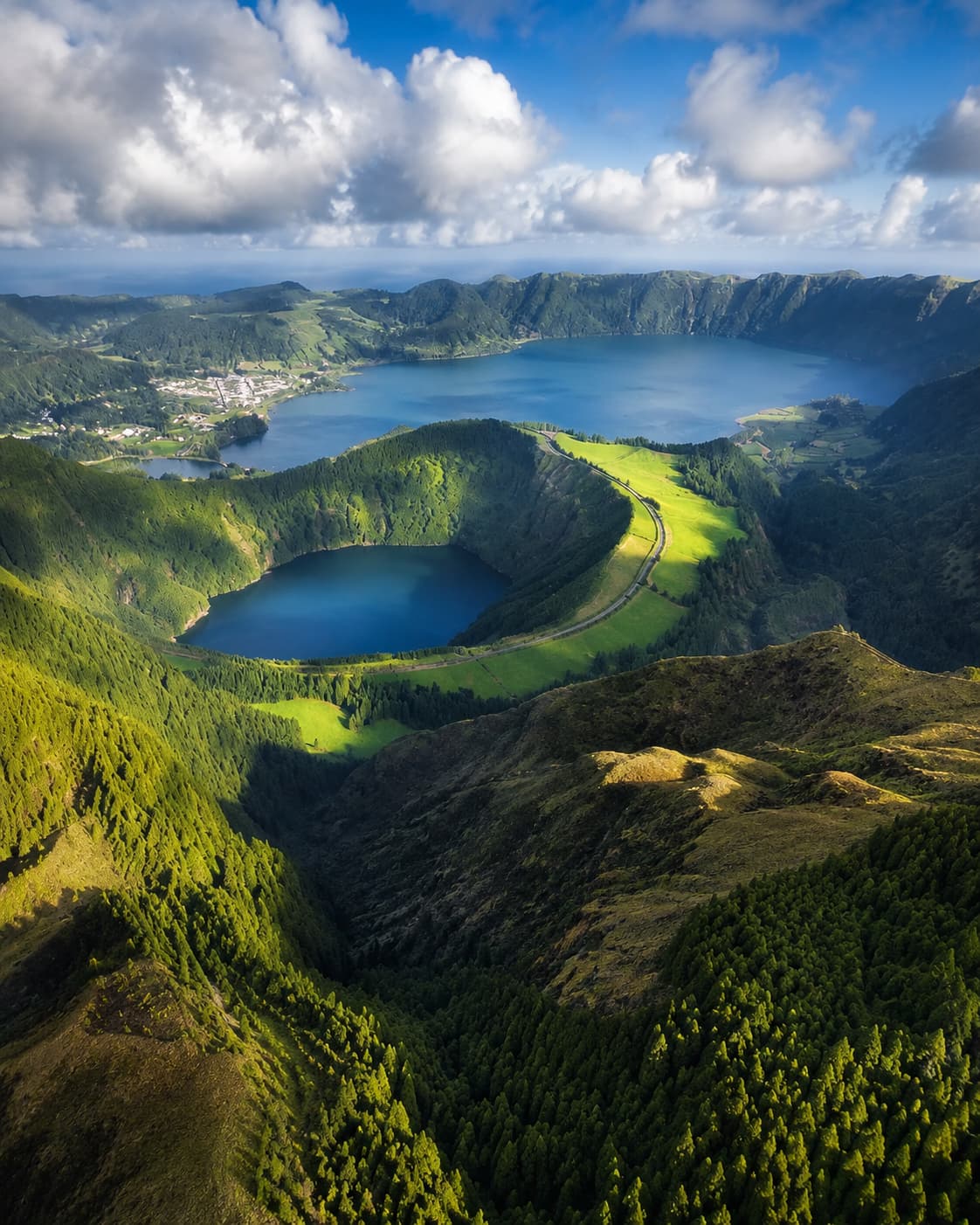 Aerial view of Sete Cidades on São Miguel, Azores — twin volcanic crater lakes of vivid blue and green surrounded by dense forest and steep caldera walls, with Atlantic clouds building overhead, showing the dramatic volcanic landscape that makes the Azores the most compelling cool-weather alternative to the Mediterranean in August.
