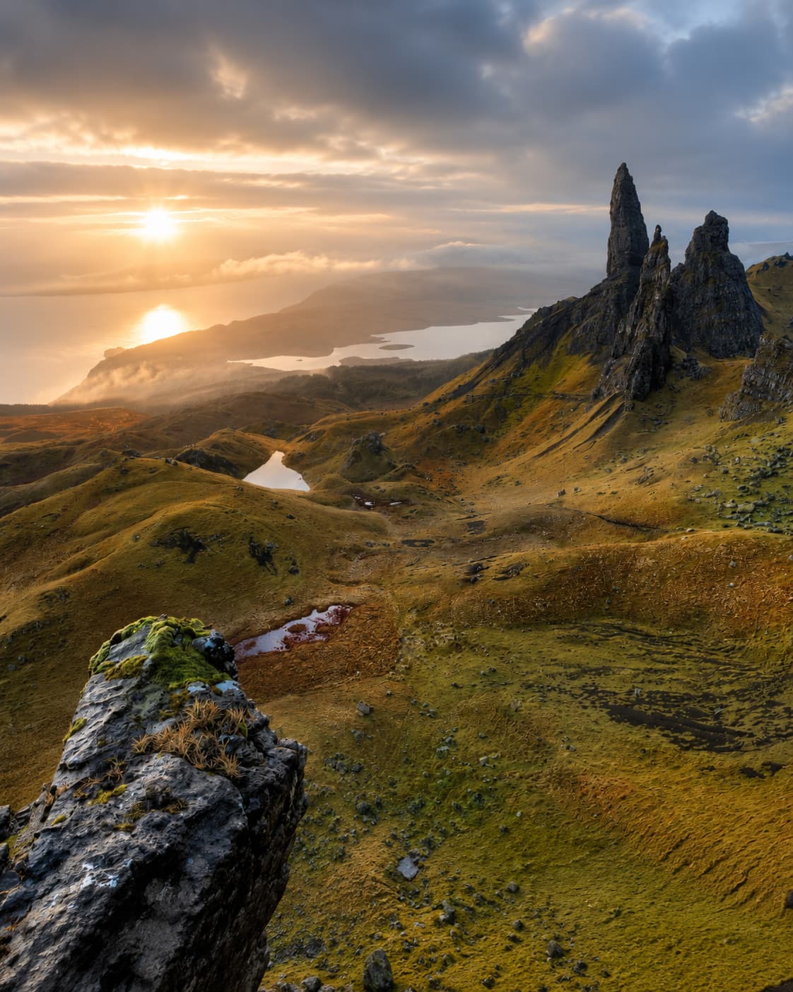 The Old Man of Storr rock formation on the Isle of Skye at dramatic sunrise — jagged black basalt pinnacles rising from rolling moorland with a loch in the valley below and golden light breaking through stormy clouds, embodying the wild, atmospheric beauty of Scotland in August.