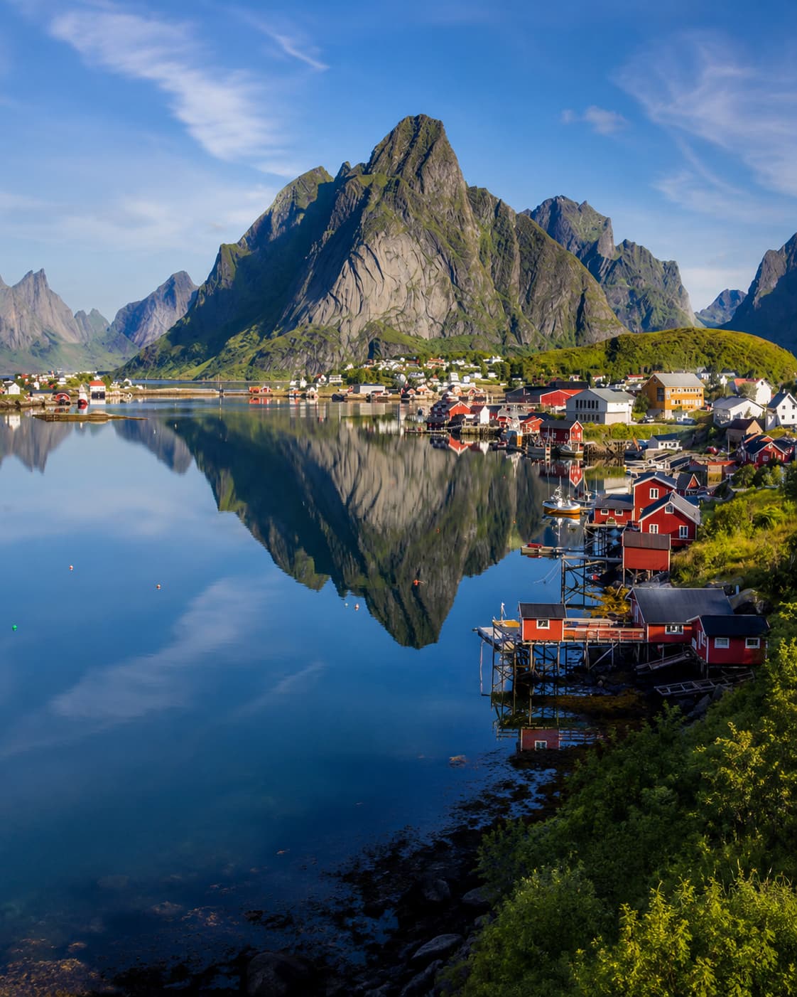 The village of Reine in the Lofoten Islands, Norway — red fishing cabins and white houses reflected perfectly in the still fjord water with dramatic granite peaks rising behind, bathed in clear summer light that shows why Lofoten is the definitive cool-weather alternative for travellers asking whether the Mediterranean is too hot in August.