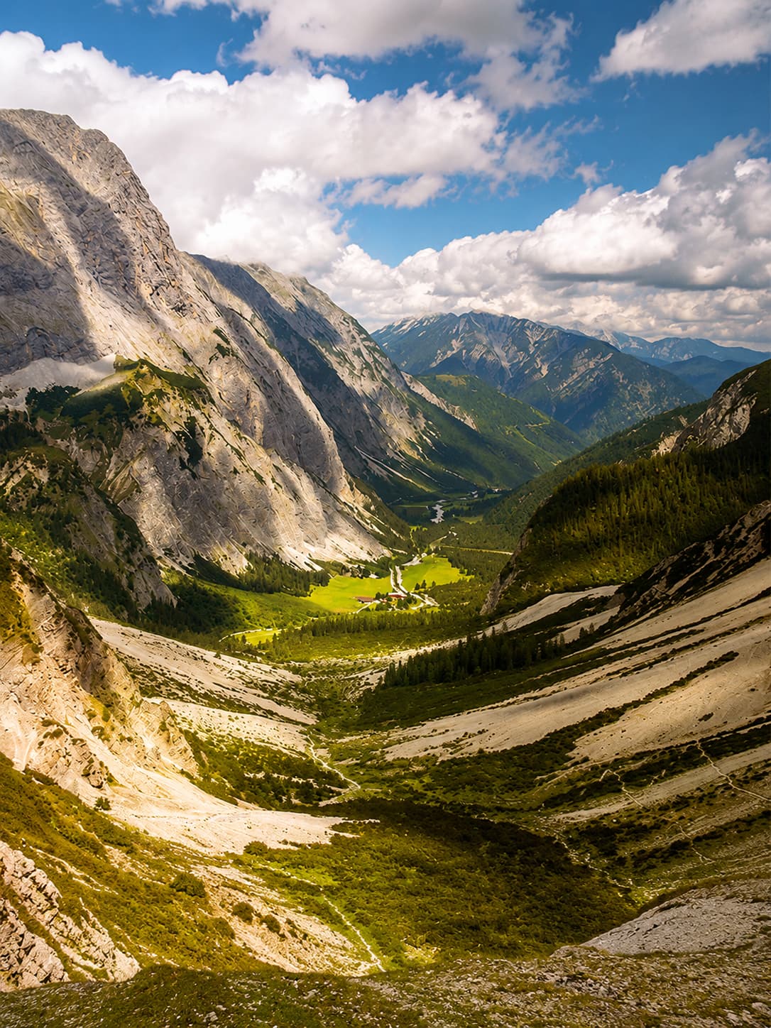 A vast Alpine valley in the Austrian or Bavarian Alps in summer — sheer limestone walls dropping to a glacially carved valley floor of vivid green meadows with a small farmstead far below, under a sky of dramatic clouds, showing the scale and cool clarity of high-altitude Europe in August.