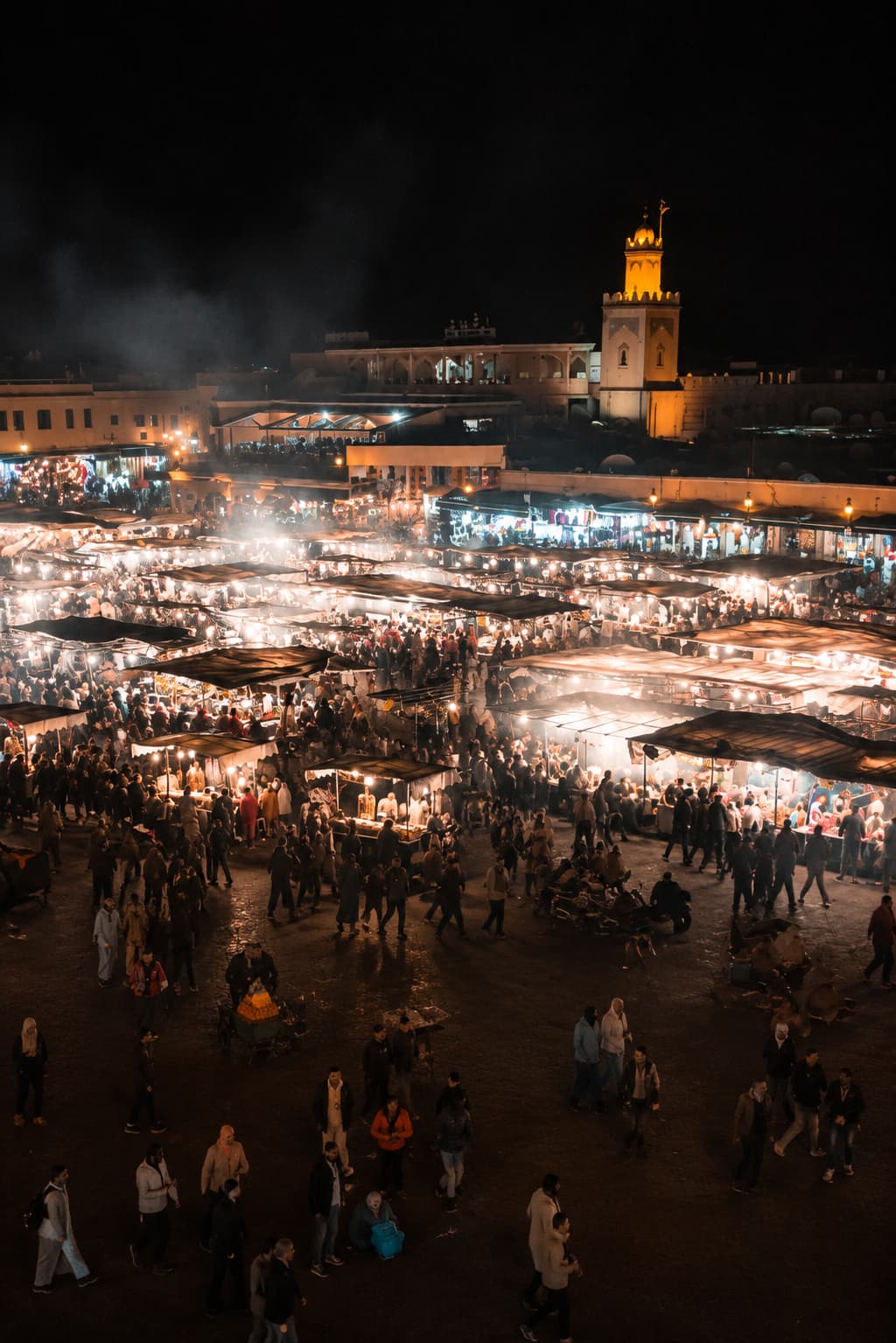 Djemaa el-Fna square in Marrakech at night — food stall lights and smoke filling the square, a mosque minaret glowing amber in the background