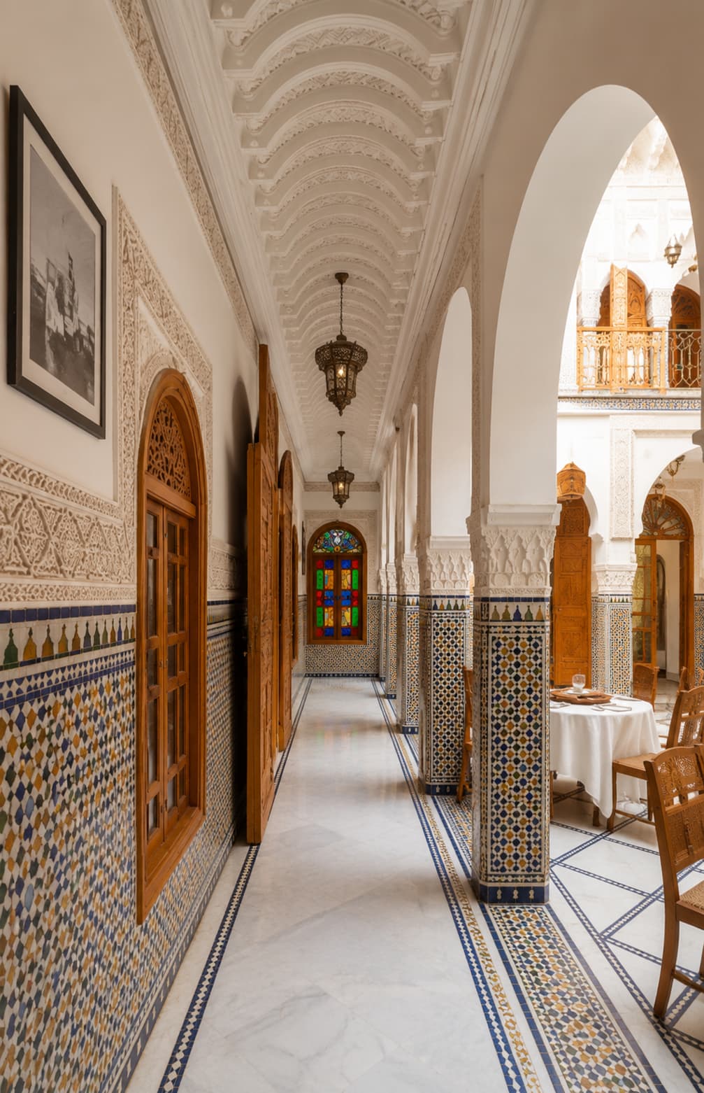 Traditional riad corridor in Morocco — zellige-tiled walls and floors, carved plasterwork ceiling, brass lanterns, and an arched view into a sunlit courtyard