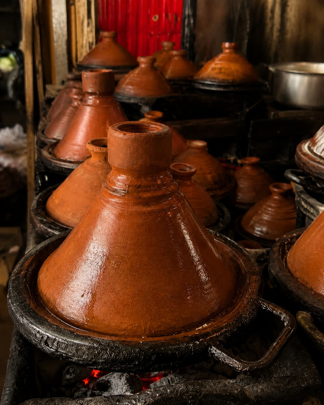 Rows of unglazed clay tagine pots with conical lids cooking over charcoal braziers in a Moroccan medina kitchen, steam visible beneath the lids