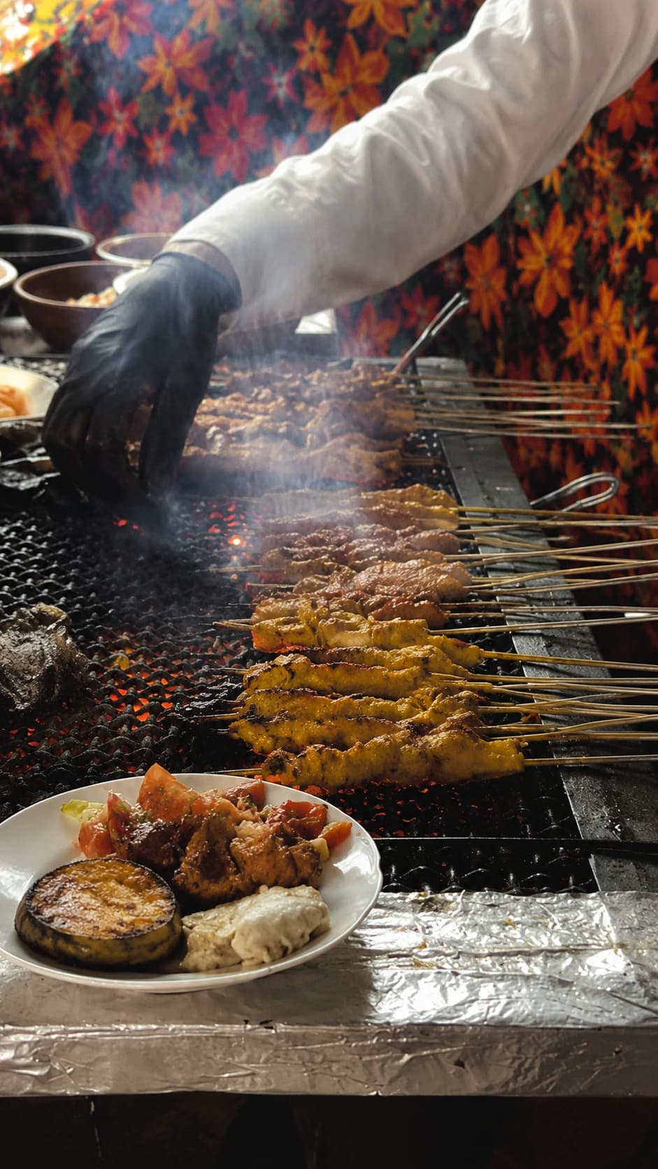 Close-up of brochette skewers grilling over red-hot charcoal at a Moroccan street food stall, with a white-gloved vendor tending the meat and a plate of salads and roasted aubergine in the foreground